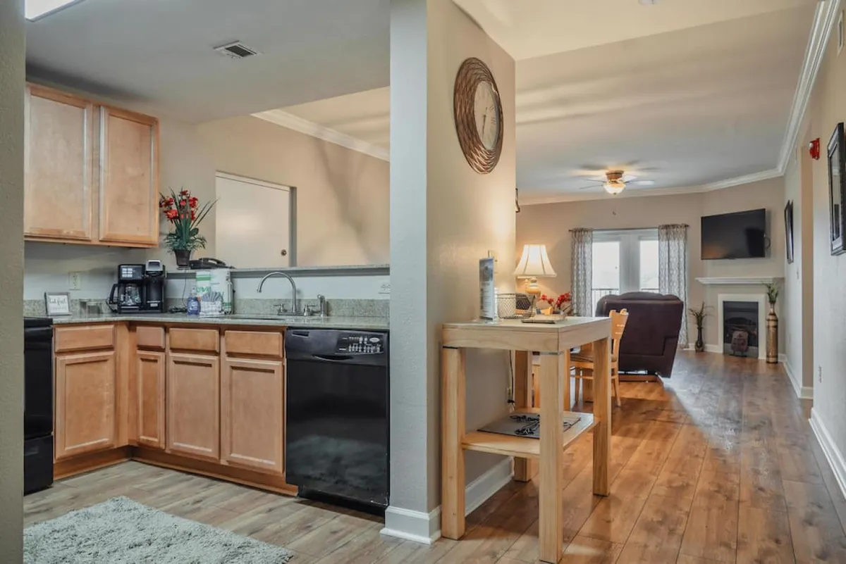 A bright, open-concept space showing the transition from the kitchen to the living area. Features include kitchen cabinetry, a dining table, and a glimpse of the living room, all styled in a welcoming and homey manner. The scene conveys seamless flow between living, dining, and kitchen areas.