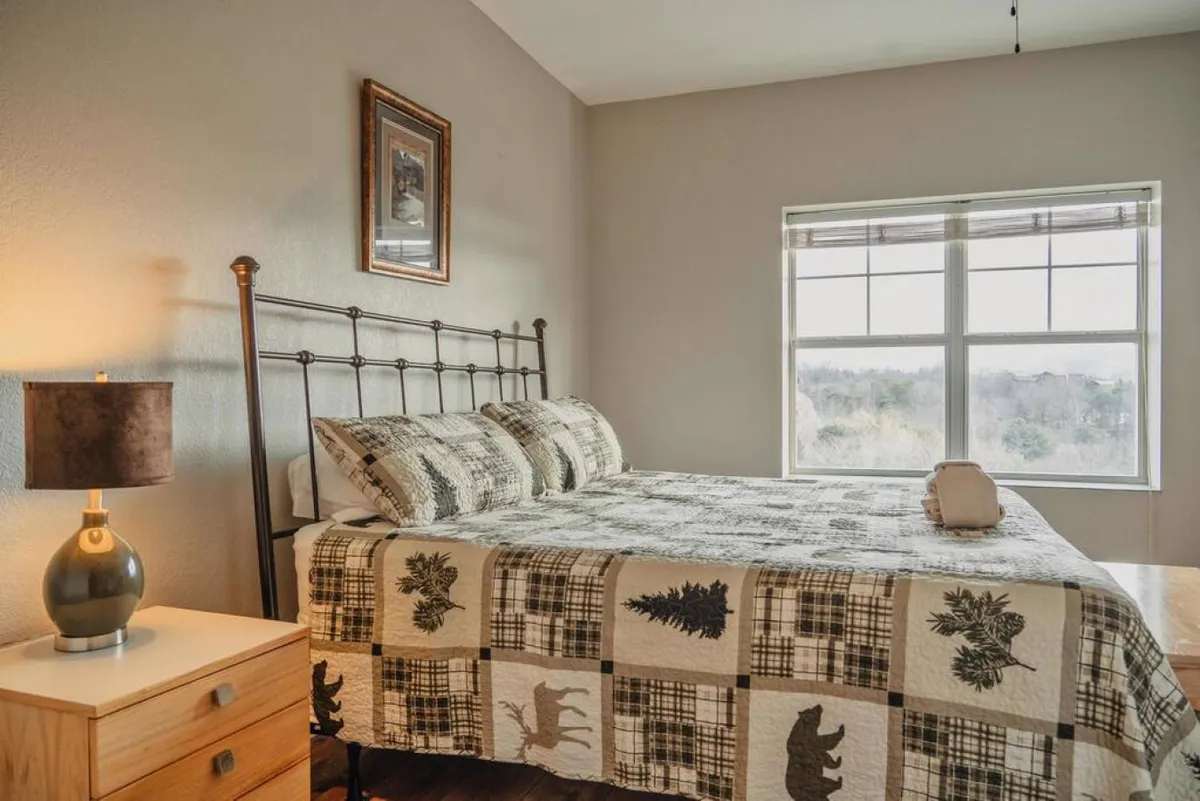 A comfortable bedroom with a patterned quilt on the bed, a bedside lamp, and a window framing beautiful nature views. The overall mood is serene and inviting, hinting at relaxation and intimate moments spent gazing outside.