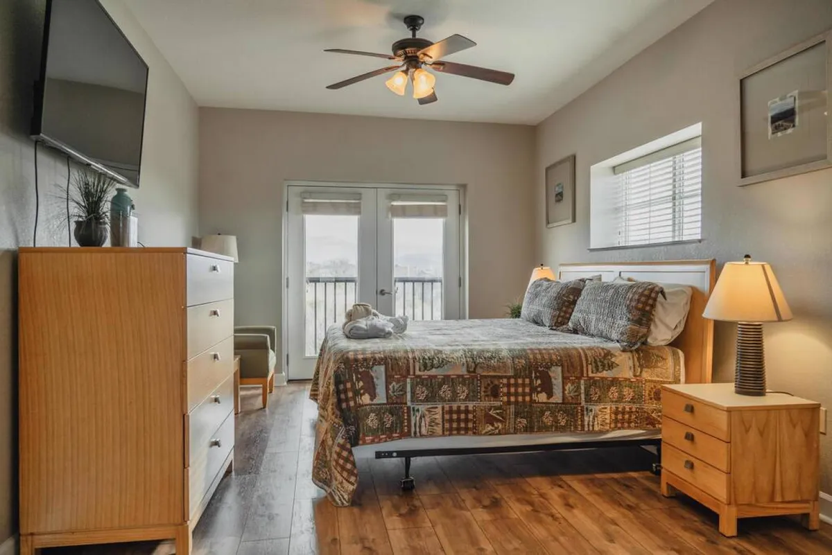 A bright, comfortable bedroom featuring a large bed with patterned bedding, expansive windows letting in natural light, a nightstand with a lamp, and a dresser. A television is visible, creating a relaxing space for both sleep and leisure.