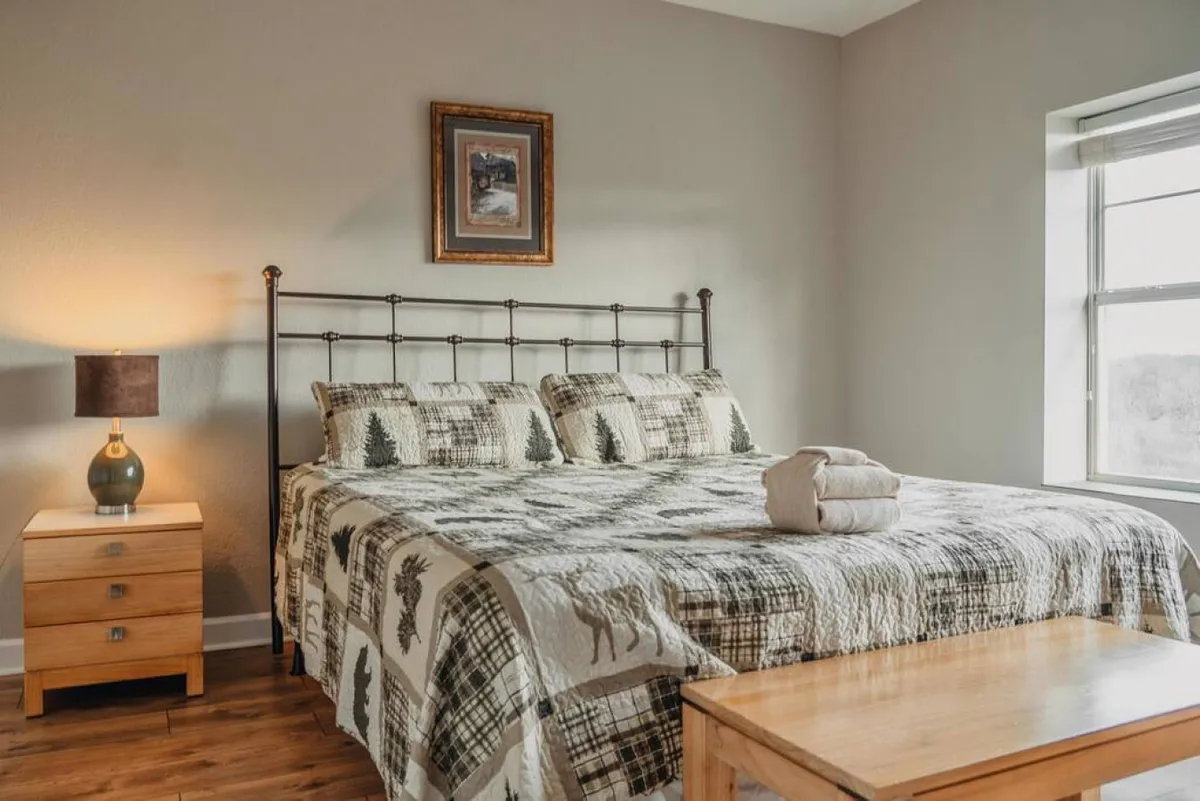 The image showcases a serene bedroom interior, featuring a comfortable bed adorned with a cozy quilt, a nightstand with a softly glowing lamp, framed artwork on the wall, and a window letting in gentle natural light. The overall mood is calm and inviting, suggesting relaxation and comfort, perfect after a day of adventure in Pigeon Forge.