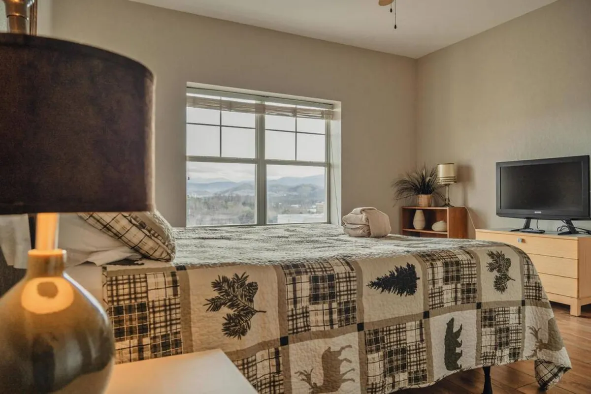 A cozy bedroom with a patterned quilt on the bed, a window offering a sweeping mountain view, and a table lamp, evoking a restful and calming atmosphere. The visible outdoor vista connects the interior comfort to the surrounding landscape.