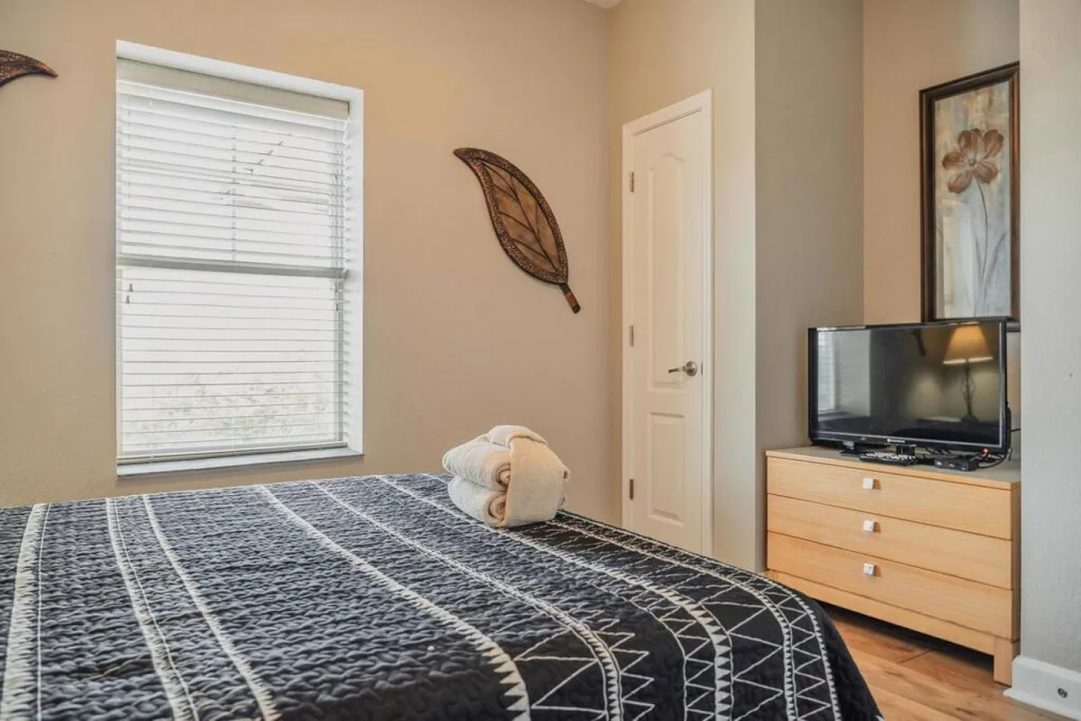 A cozy bedroom with a bed covered with a patterned quilt, a television set atop a dresser, towels folded on the bed, a window with blinds, and decorative wall art, creating a homey and restful atmosphere.