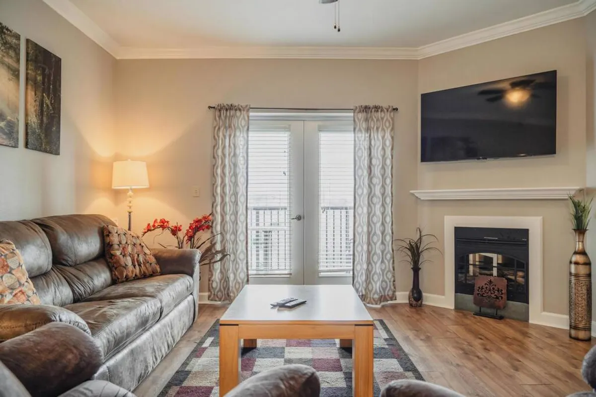 A spacious, cozy living room featuring a comfortable sofa, coffee table, a large flat screen TV, and a fireplace. Generous windows, framed by curtains, allow natural light to filter in, highlighting both the plush seating and the inviting, communal atmosphere of the space.