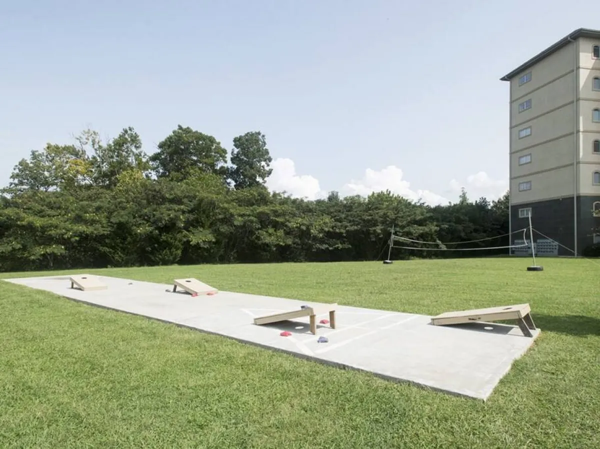 A vibrant exterior view featuring cornhole boards set up on a lush grass lawn, framed by tall trees and the building in the background. The setup suggests space for group activities and casual social gatherings outdoors.