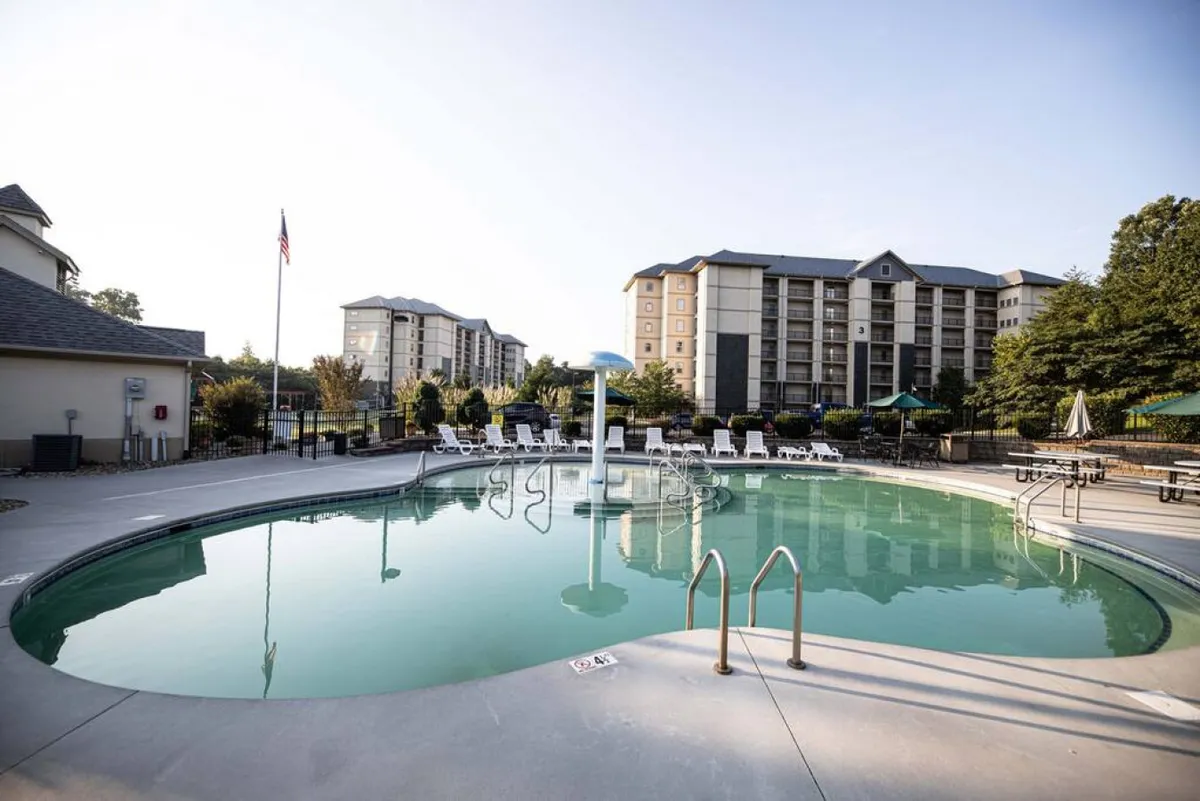 This image showcases an inviting outdoor swimming pool surrounded by loungers and beautifully landscaped greenery, with resort buildings visible in the background. Children and families can be imagined splashing and playing, while adults relax nearby.