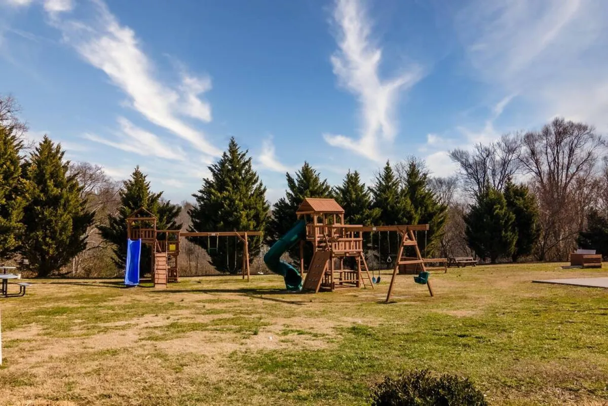 A colorful playground structure with swings and a slide sits beside a shaded picnic area. Families and children are engaged in playful activities, while others enjoy a meal outdoors, surrounded by green space.