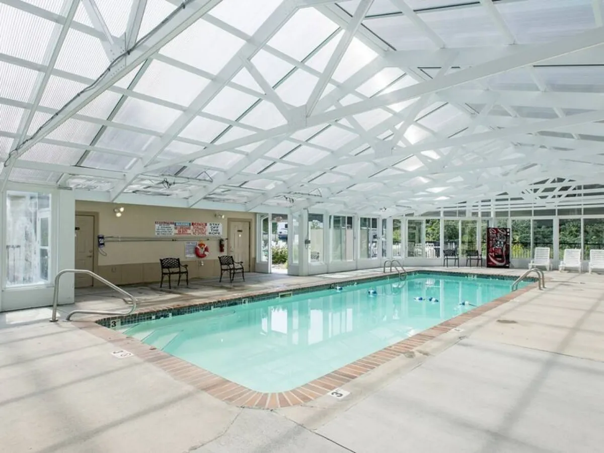 A bright indoor swimming pool with a glass ceiling, white seating areas, and plenty of natural light streaming in. The atmosphere is airy and inviting, perfect for both relaxing swims and lively family gatherings, regardless of the season.